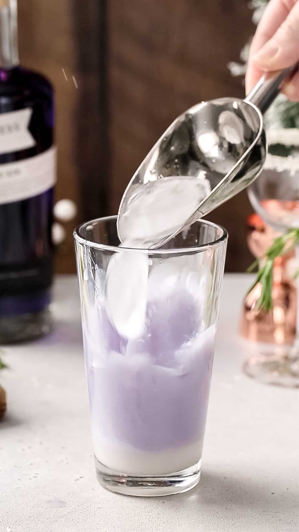 Hand adding ice to a cocktail shaker filled with purple liquid.