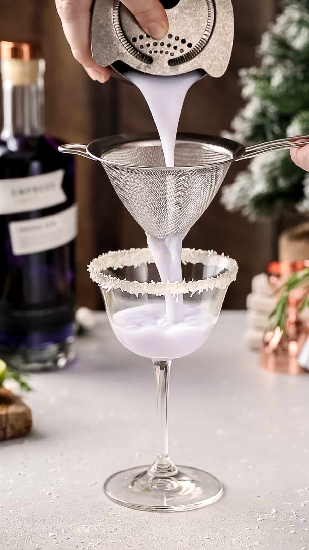 Hand double straining a purple cocktail into a coupe glass with a coconut rim.