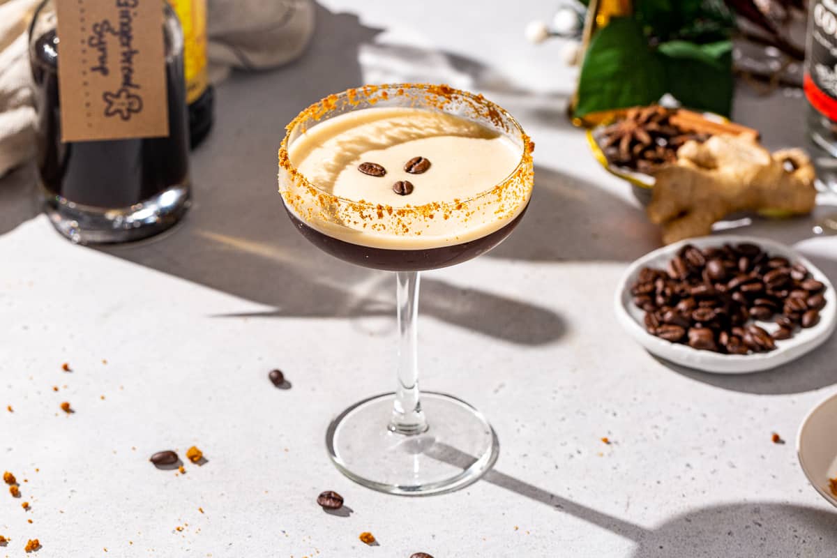 Gingerbread Espresso Martini in a stemmed cocktail glass on a countertop. In the background are coffee beans, gingerbread syrup and vodka along with fresh ginger and some Christmas decor.
