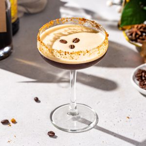 Gingerbread Espresso Martini in a stemmed cocktail glass on a countertop. The drink is brown with a foam layer on top, and it has cookie crumbs on the rim along with three coffee beans floating on top.