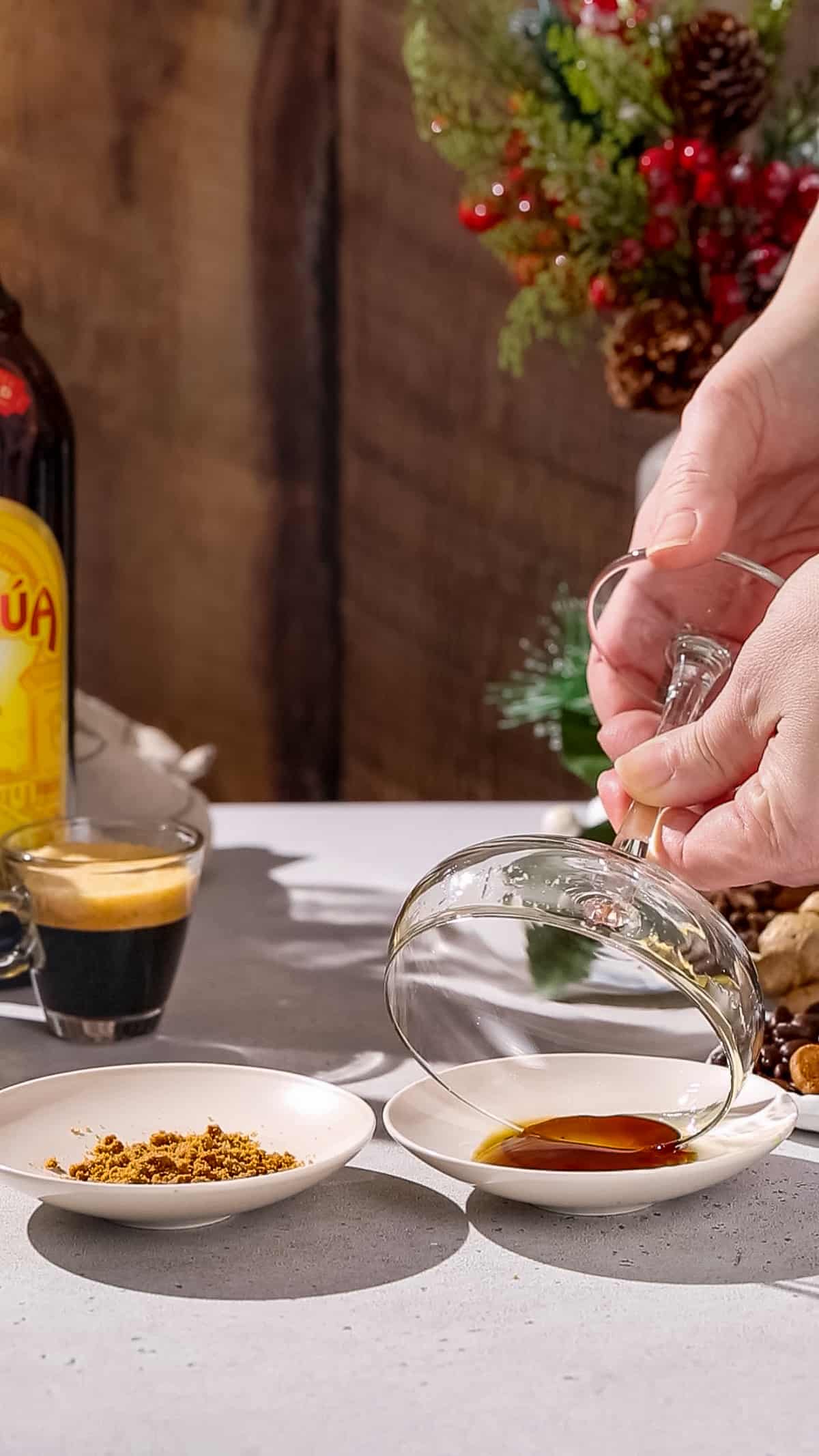 Hands dipping the rim of a coupe glass into a dish of gingerbread syrup.