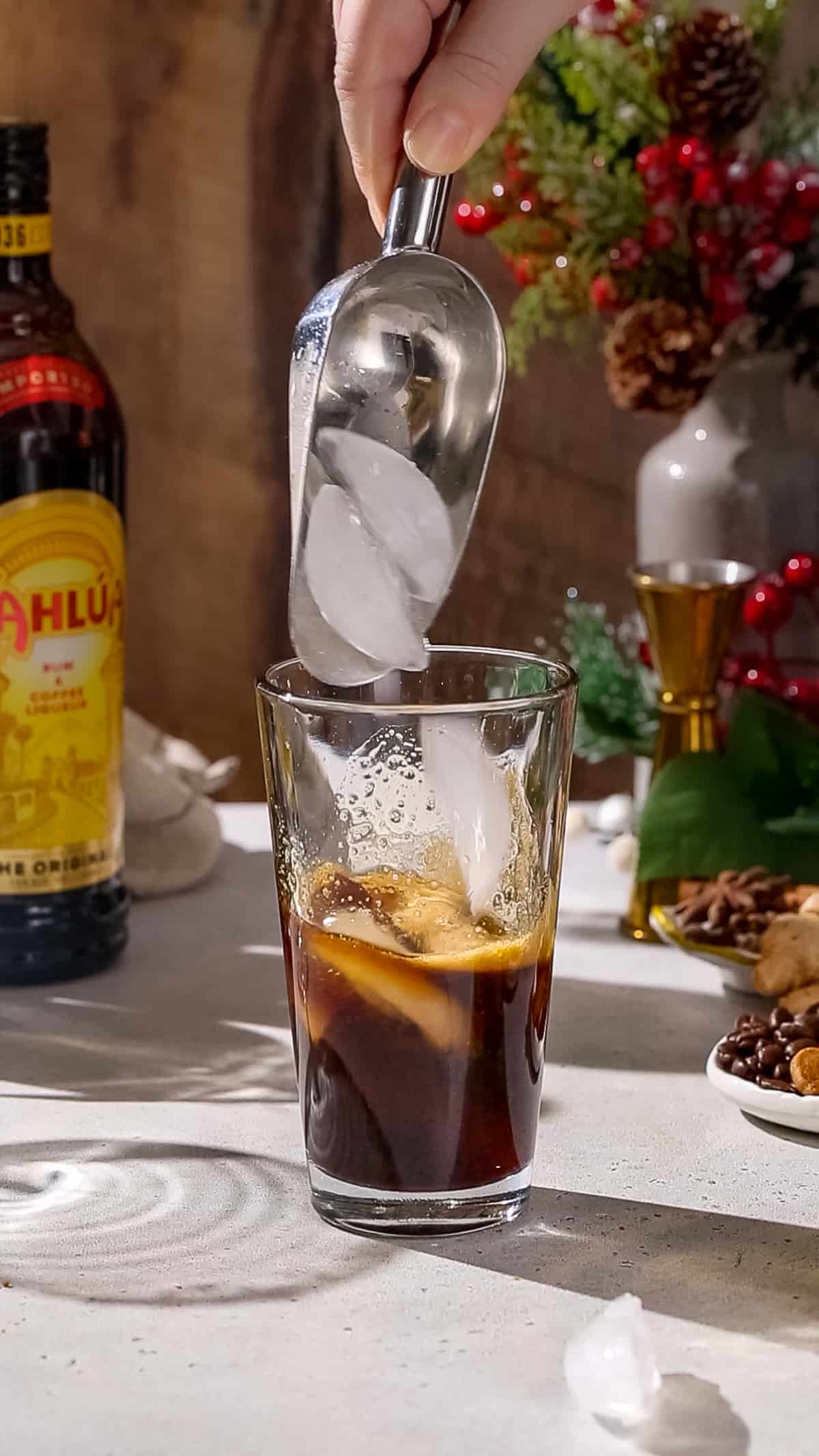 Hand using an ice scoop to add ice to a cocktaiil shaker filled with brown liquid.