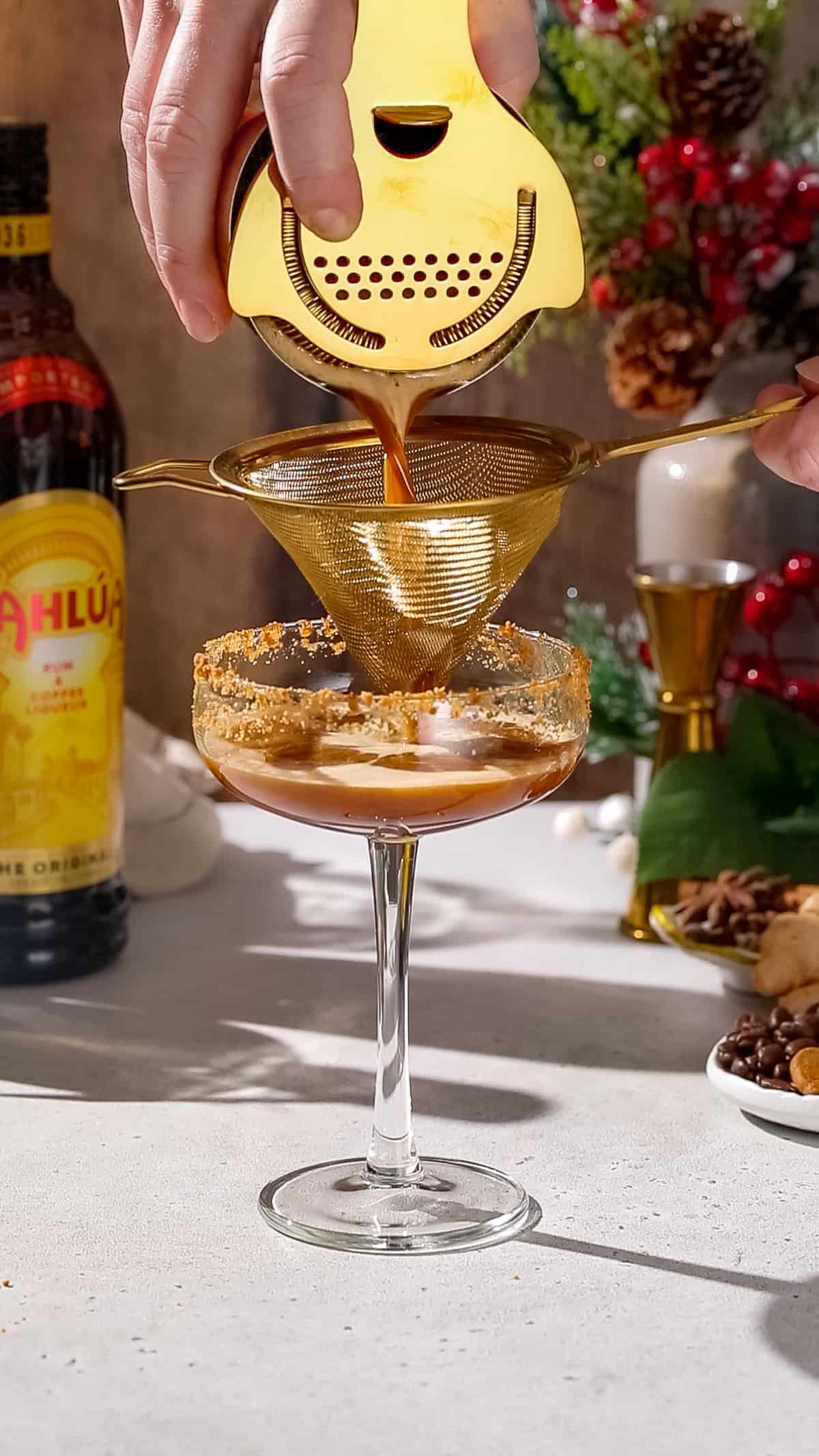 Hands double straining a cocktail into a glass with a cookie crumb rim.