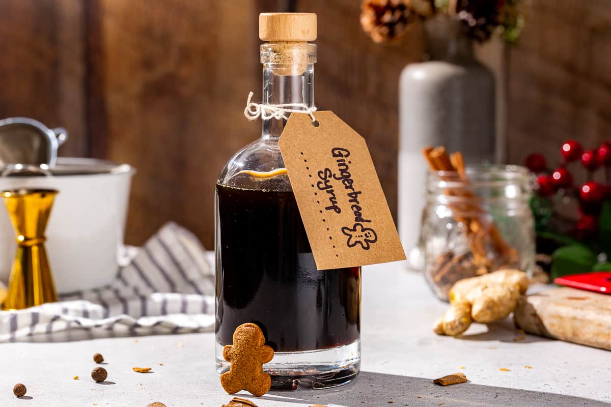 A glass bottle of Gingerbread syrup on a countertop with a kraft paper label that says “Gingerbread Syrup” with a hand drawn gingerbread man. Some fresh ginger and cinnamon sticks are seen in the background. A mini gingerbread man is resting against the bottle.
