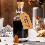 A glass bottle of Gingerbread syrup on a countertop with a kraft paper label that says “Gingerbread Syrup” with a hand drawn gingerbread man. Some fresh ginger and cinnamon sticks are seen in the background. A mini gingerbread man is resting against the bottle.