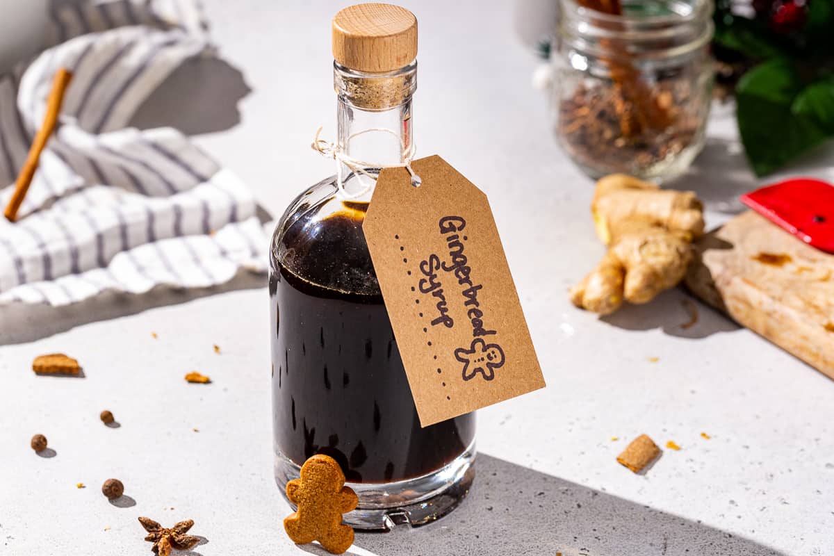 A glass bottle of Gingerbread syrup on a countertop with a kraft paper label that says “Gingerbread Syrup” with a hand drawn gingerbread man. Some fresh ginger and cinnamon sticks are seen in the background. A mini gingerbread man is resting against the bottle.