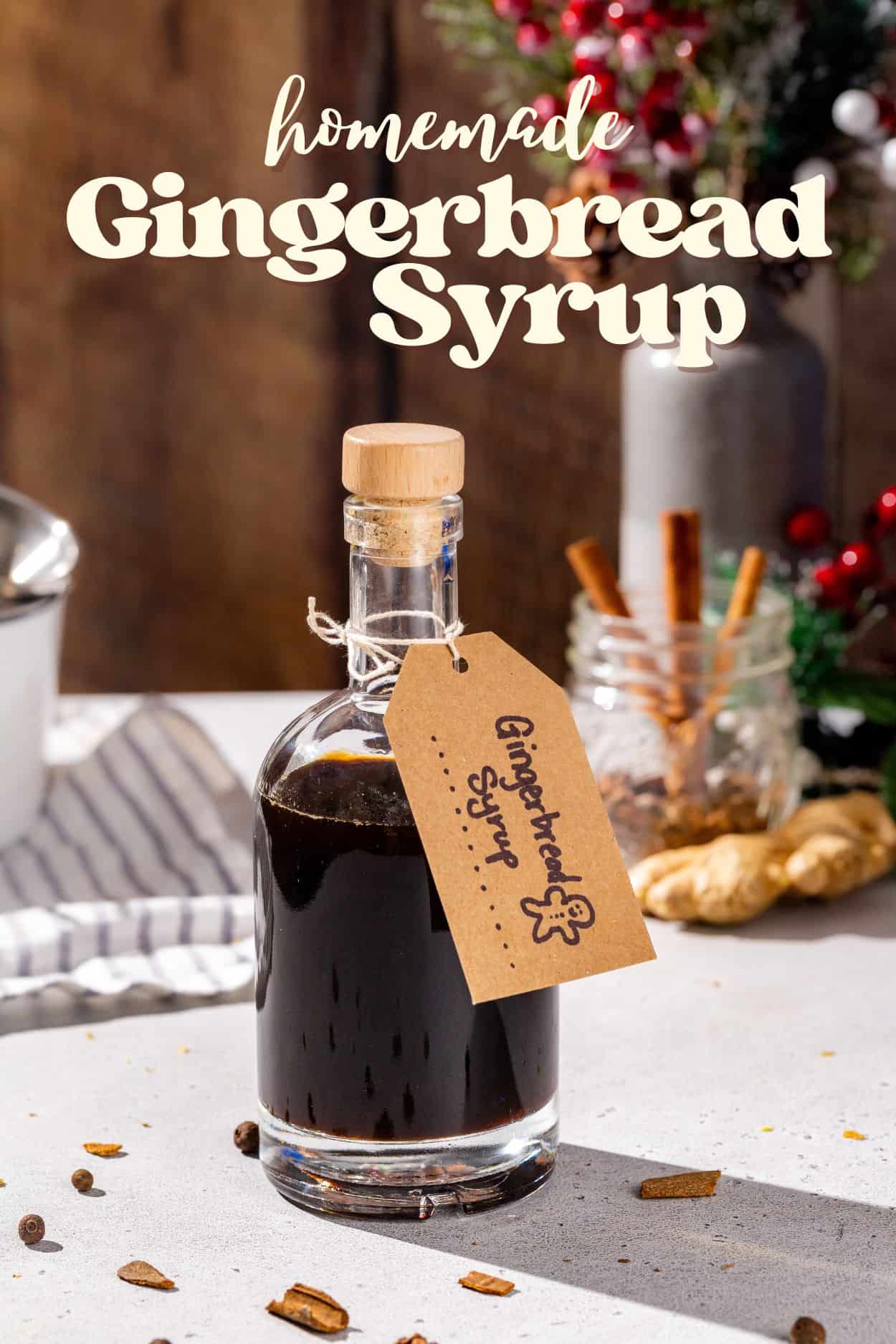 A glass bottle of Gingerbread syrup on a countertop with a kraft paper label that says “Gingerbread Syrup” with a hand drawn gingerbread man. Ingredients and equipment are in the background. Text overlay above the bottle says “Homemade Gingerbread Syrup”.