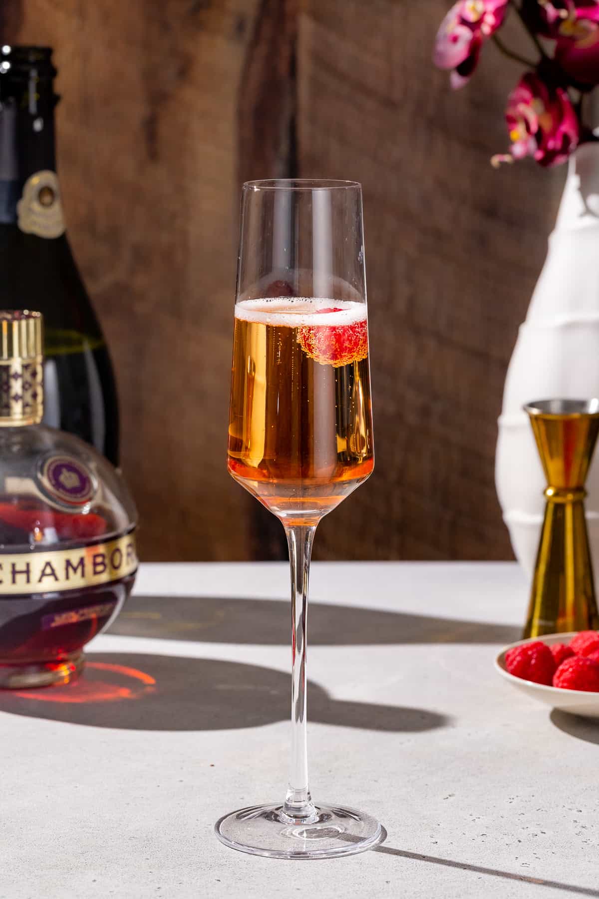 Side view of a Kir Imperial cocktail in a champagne flute. The drink is light orange and has bubbles at the top, and a single raspberry is floating in the glass. Bottles of Chambord and Champagne are in the background along with raspberries and a gold jigger.