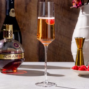 Side view of a Kir Imperial cocktail in a champagne flute. A raspberry is in the drink as a garnish. Bottles of Chambord and Champagne are in the background along with fresh raspberries and a gold jigger.