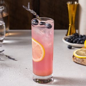 Side view of a Blueberry Lavender Lemonade mocktail. The drink is pink colored and garnished with blueberries, lavender and a lemon slice. In the background are some ingredients and bar tools.