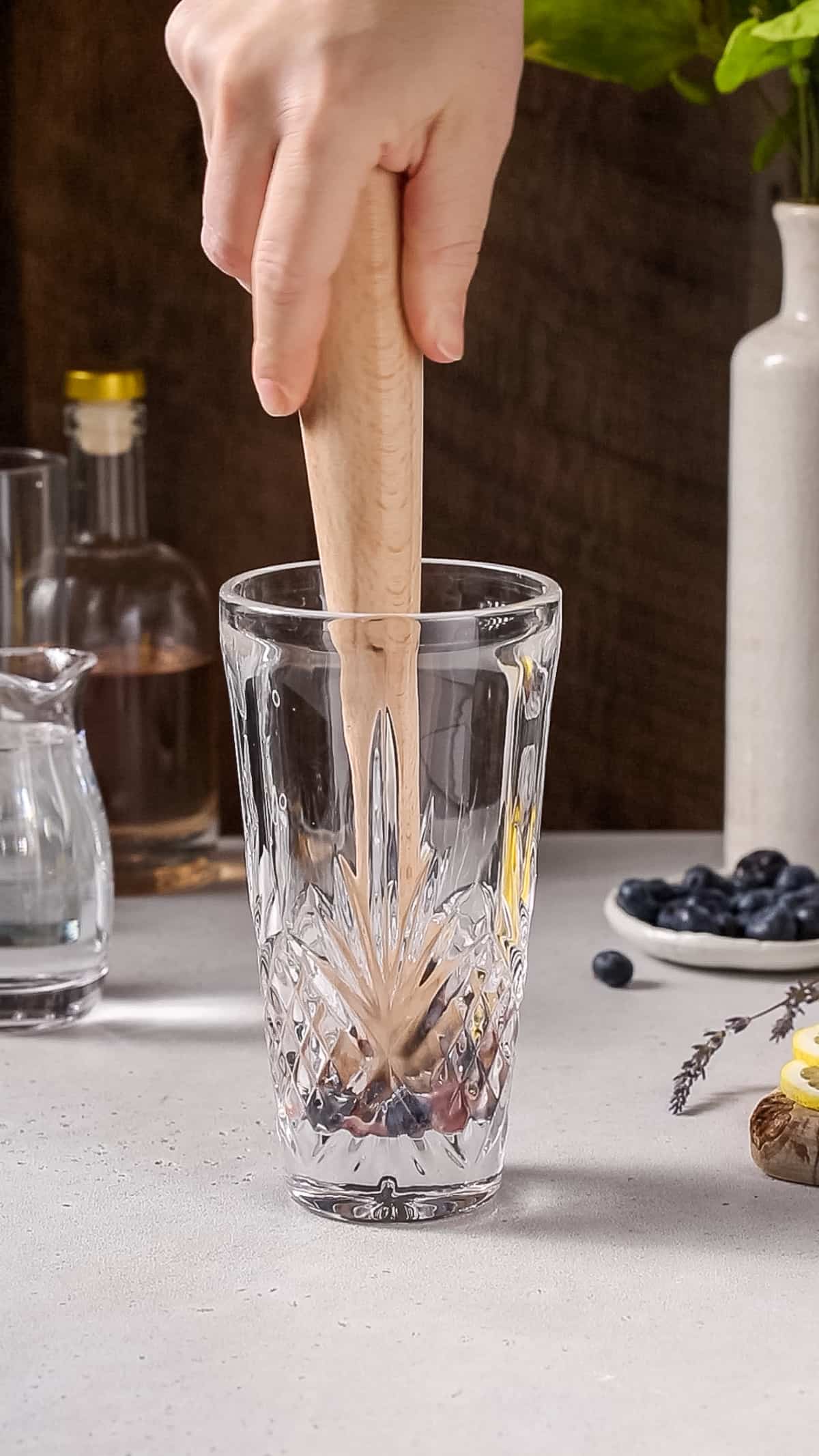 Hand using a wooden mudder to muddle blueberries in a cocktail shaker.