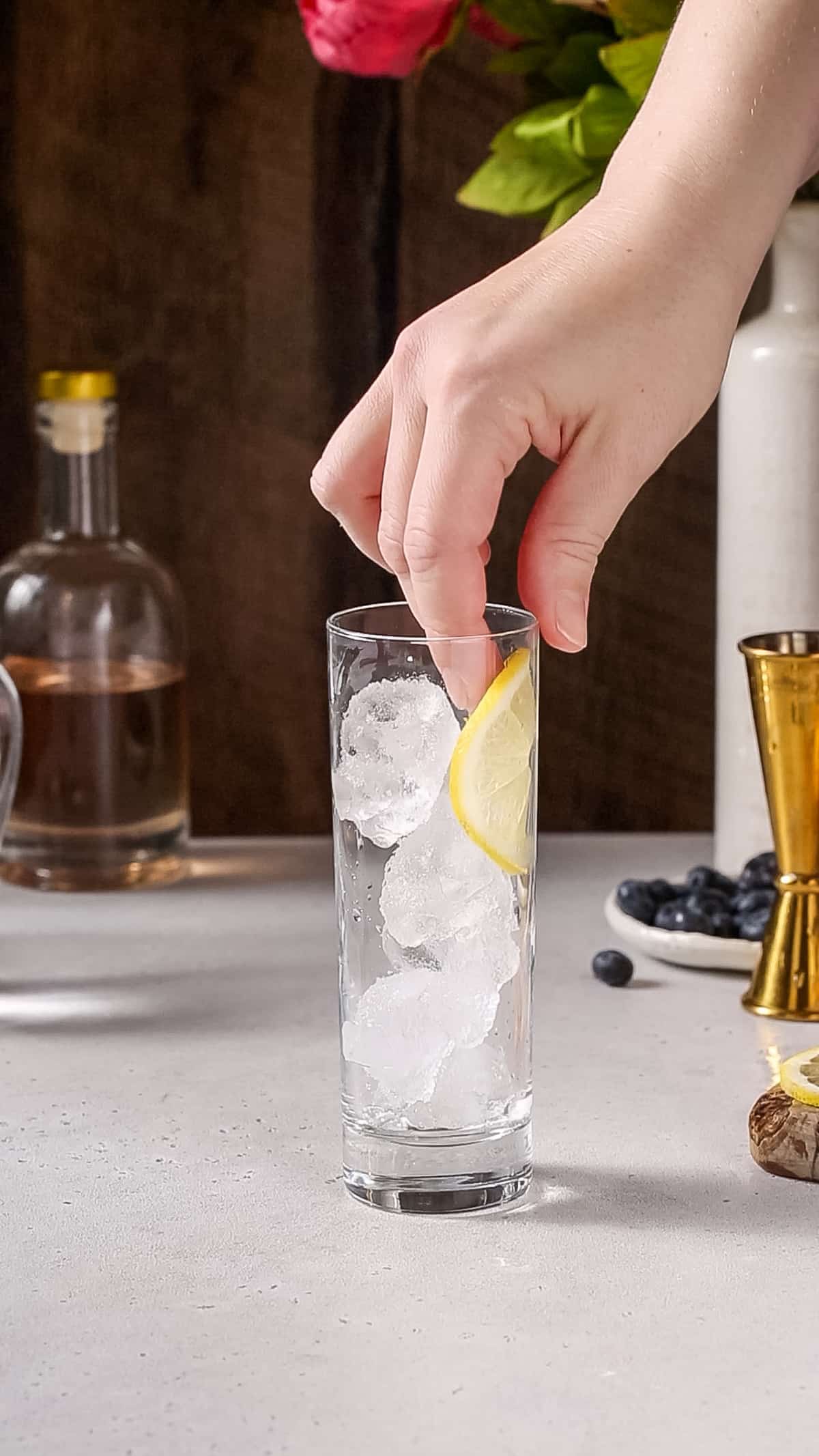 Hand adding a lemon slice to the side of a collins glass filled with ice.