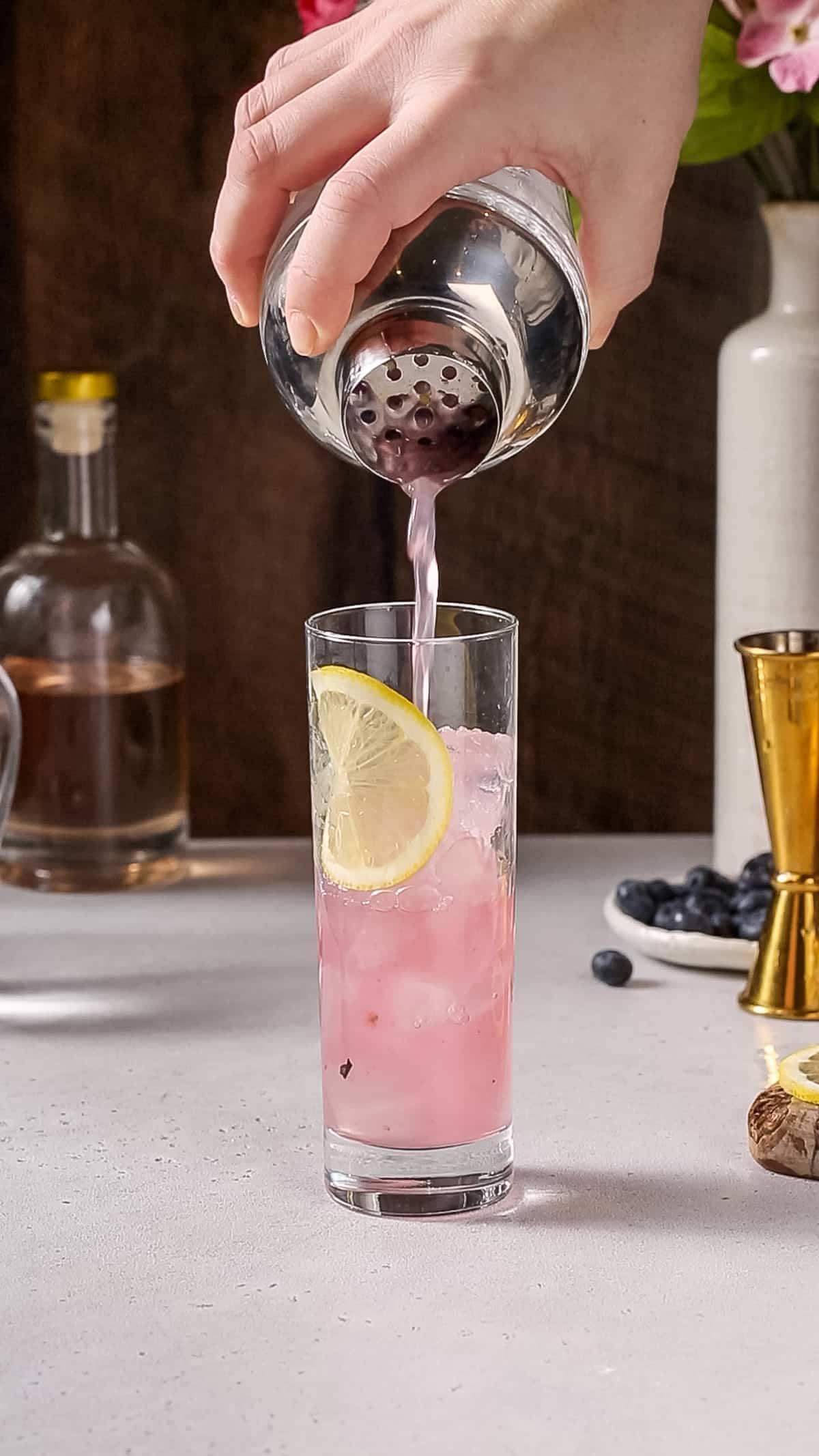 Hand straining a pink liquid from a cocktail shaker into a prepared collins glass.