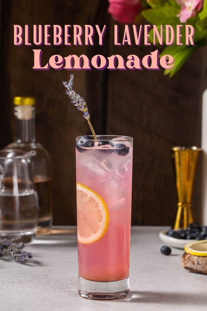 Side view of a Blueberry Lavender Lemonade mocktail. The drink is pink colored and garnished with blueberries, lavender and a lemon slice. In the background are some ingredients and bar tools. Text overlay at the top says "Blueberry Lavender Lemonade" in bold pink letters.