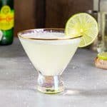 Coconut lime mocktail in a martini glass. The drink has a lime slice garnish. Topo Chico seltzer water and some other ingredients are in the background.