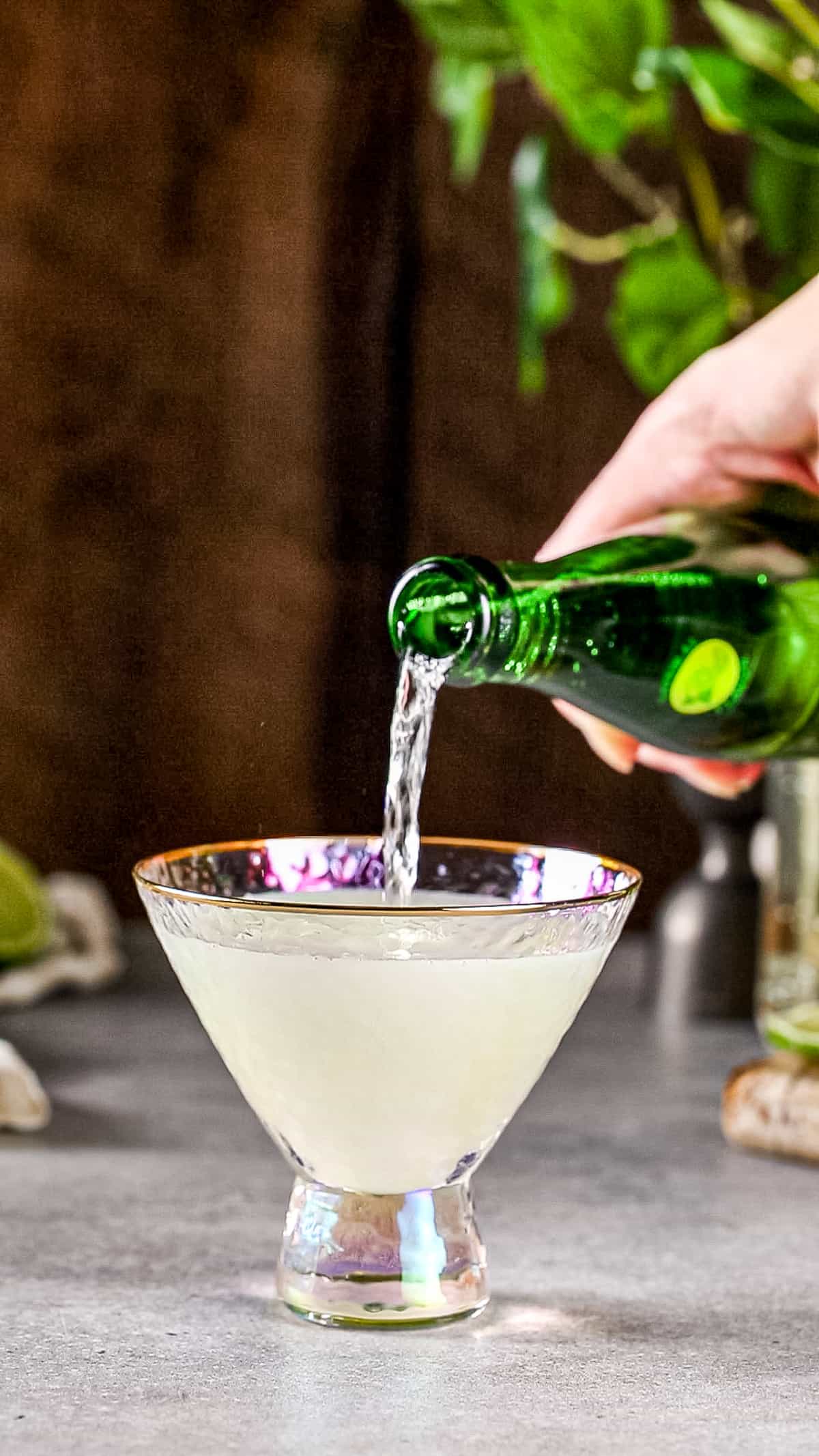 Hand pouring sparkling water into a martini glass filled with liquid.