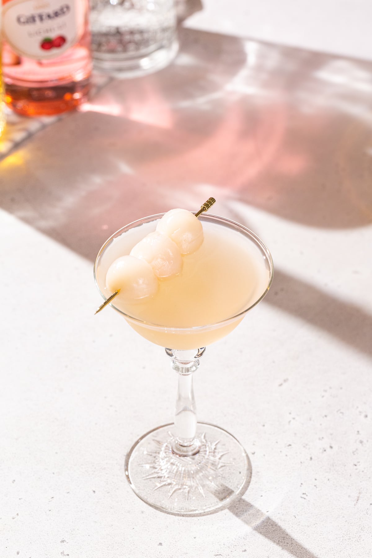 Slightly overhead view of a Lychee gin martini in a coupe glass. In the background you can see part of a bottle of gin and a bottle of lychee liqueur.