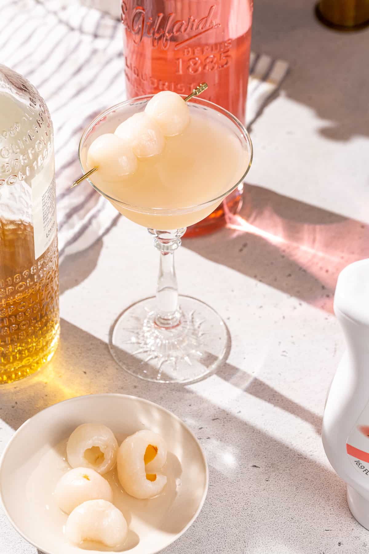 Lychee Gin martini on a countertop. The drink is in a coupe glass and is surrounded by lychee fruits, lychee puree, vermouth and lychee liqueur.