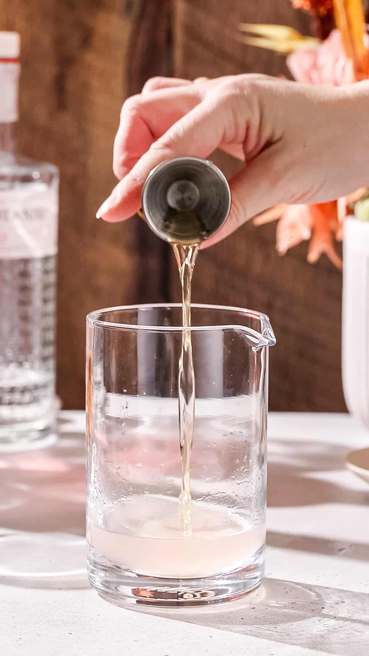 Hand pouring dry vermouth into a cocktail mixing glass.