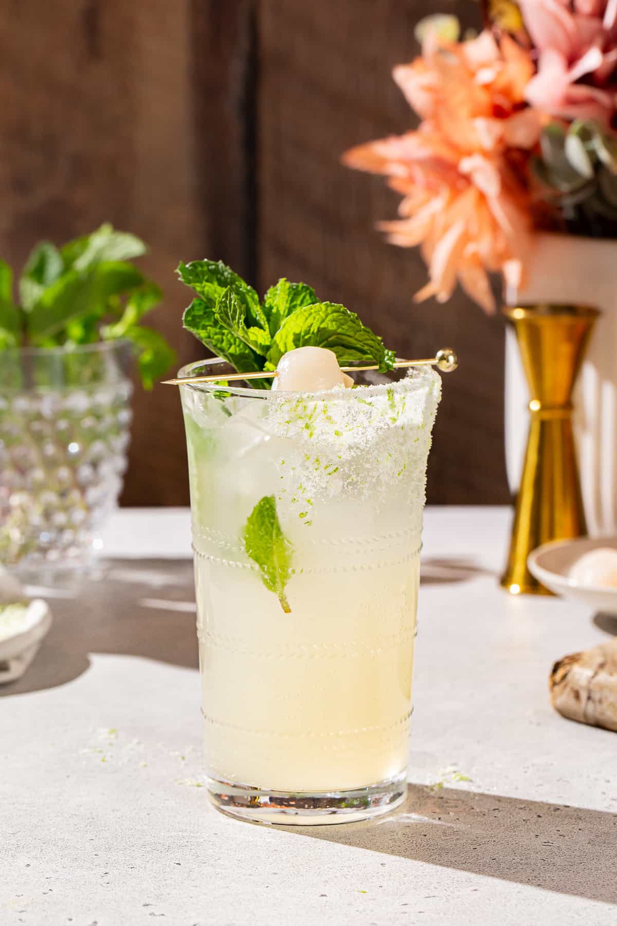 Side view of a Lychee mocktail garnished with mint and a lychee along with lime zest sugar on the side of the glass. In the background are flowers, a jigger, a dish of lychees and fresh mint.