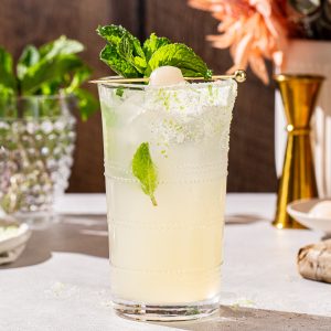 Side view of a Lychee mocktail garnished with mint and a lychee along with lime zest sugar on the side of the glass. In the background are flowers, a jigger, a dish of lychees and fresh mint.