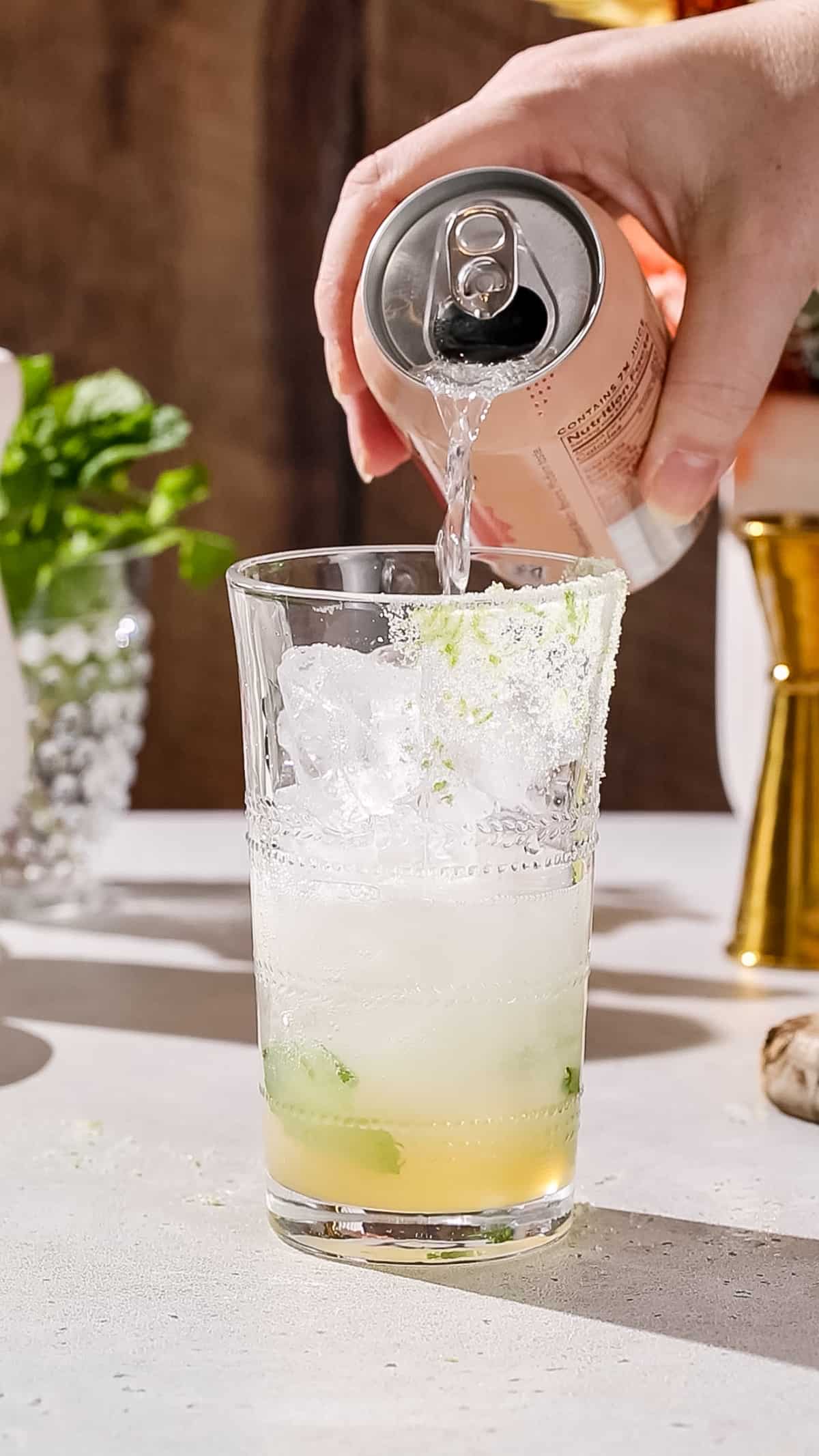 Hand pouring lychee flavored seltzer into a cocktail glass filled with ice and mint leaves.