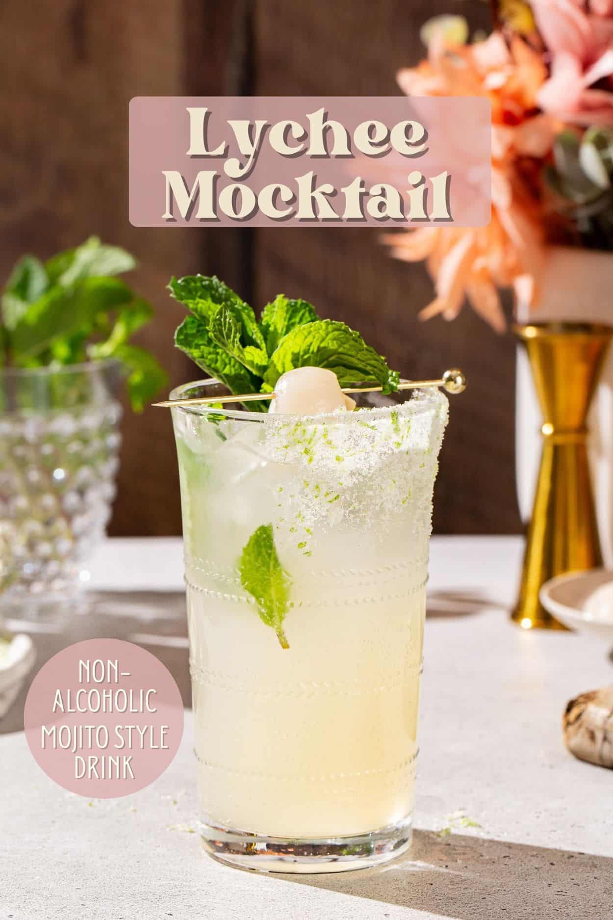 Lychee Mocktail on a countertop with bar tools and ingredients in the background. The drink is in a tall glass and garnished with a whole lychee and mint. Text overlay at the top says “Lychee Mocktail” and at the bottom left says “Non-Alcoholic Mojito Style Drink”.