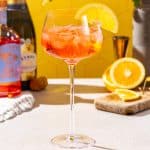 Side view of a Non-alcoholic Aperol Spritz drink on a countertop. A cut orange is in the background along with the Lyre’s Italian Spritz and Lyre’s Classico bottles.