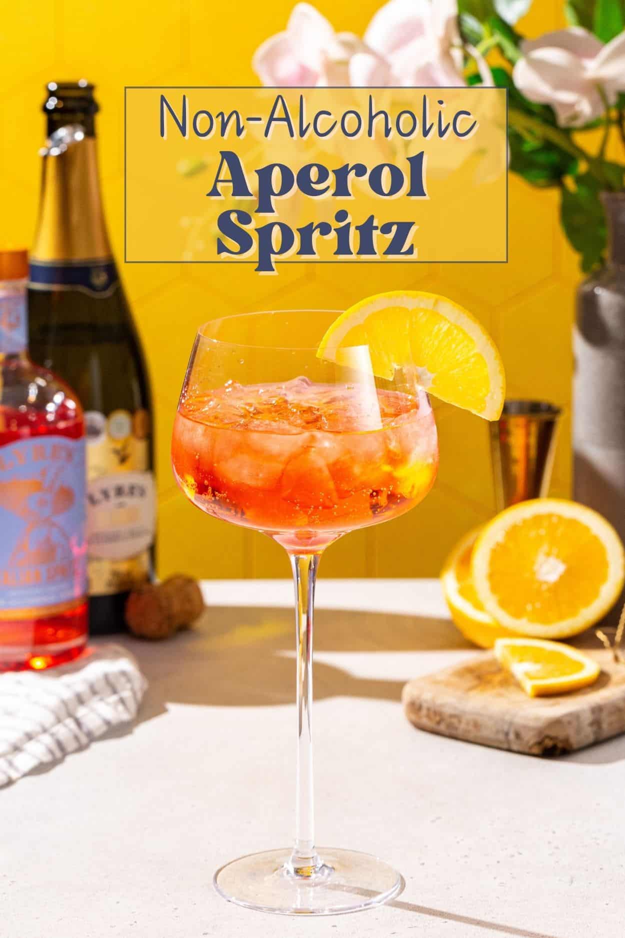 Non-alcoholic Aperol Spritz mocktail on a countertop. In the background are ingredients and flowers invase. Text overlay above the drink says “Non-Alcoholic Aperol Spritz”.