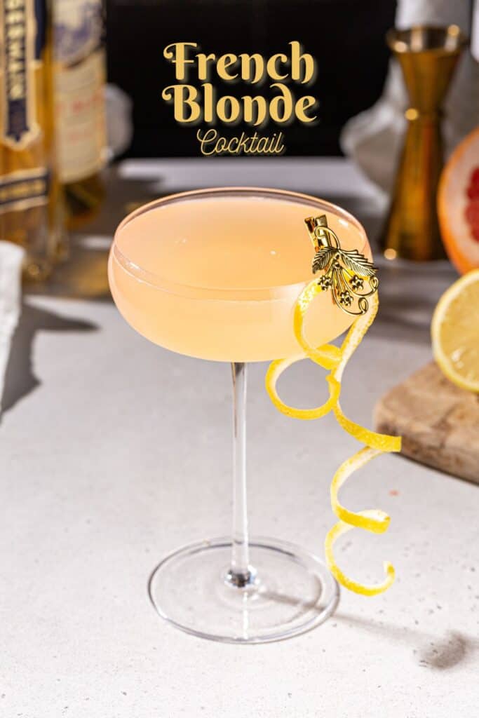 French Blonde cocktail in a coupe glass on a countertop. The drink is garnished with a long curly lemon peel held onto the glass with a gold clip. In the background are some ingredients used to make the drink. Text overlay at the top says “French Blonde cocktail”.
