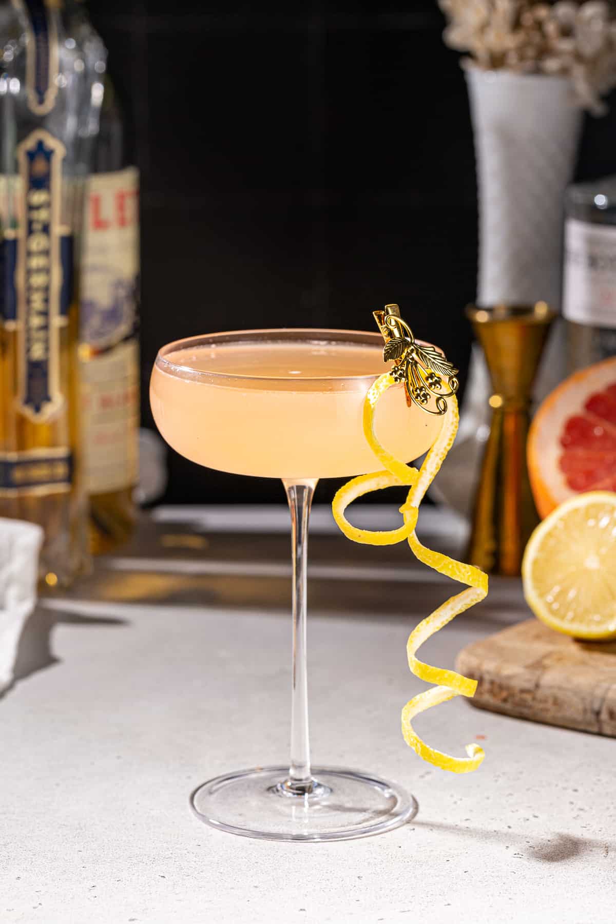 Side view of a French Blonde cocktail in a coupe glass with a curly lemon peel garnish. There are ingredients and bar tools in the background.