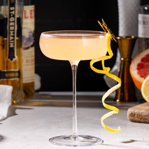 Side view of a French Blonde cocktail with a curly lemon peel garnish. In the background are bottles of St Germain, Lillet Blanc, and gin along with a grapefruit, lemon, and a gold jigger.