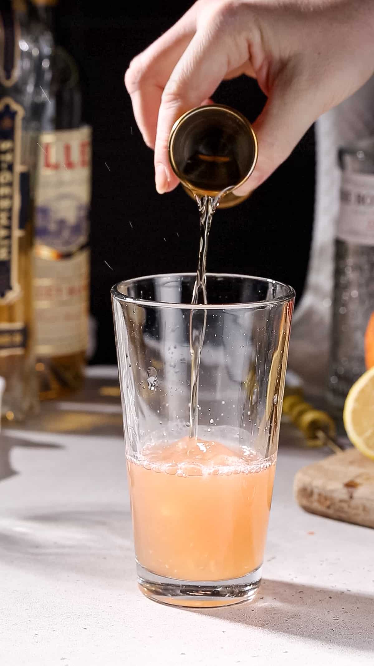 Hand pouring elderflower liqueur into a cocktail shaker.