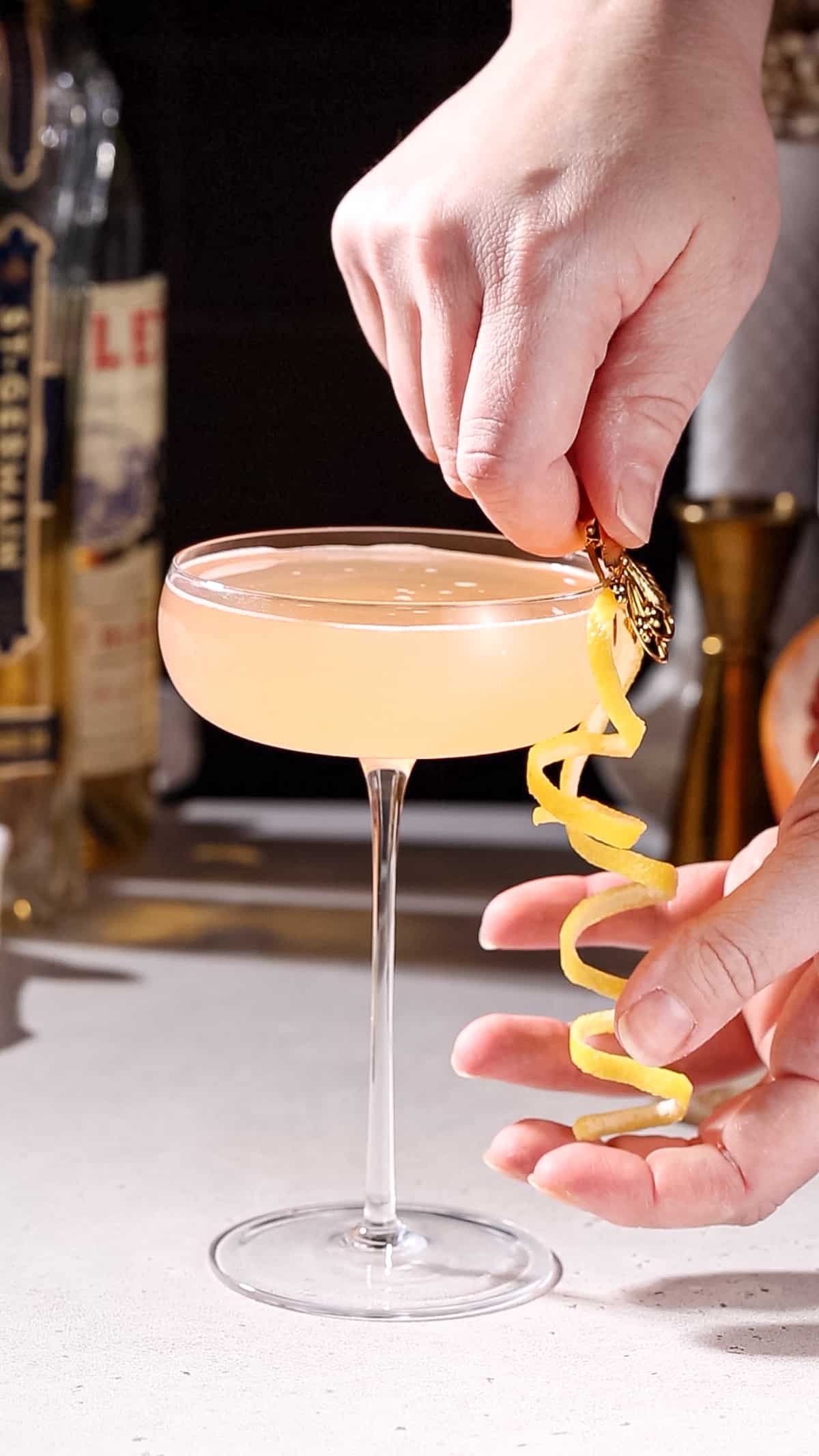 Hand adding a long curly lemon peel garnish to a coupe glass using a clip.