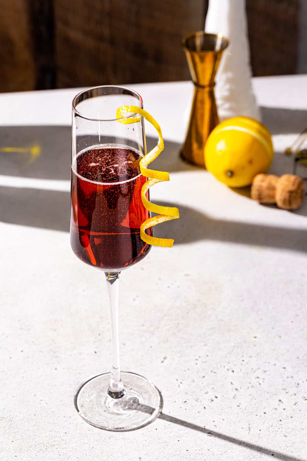 Kir Royale cocktail in a champagne flute with lemon peel garnish. A lemon along with a gold jigger and a champagne cork are in the background.