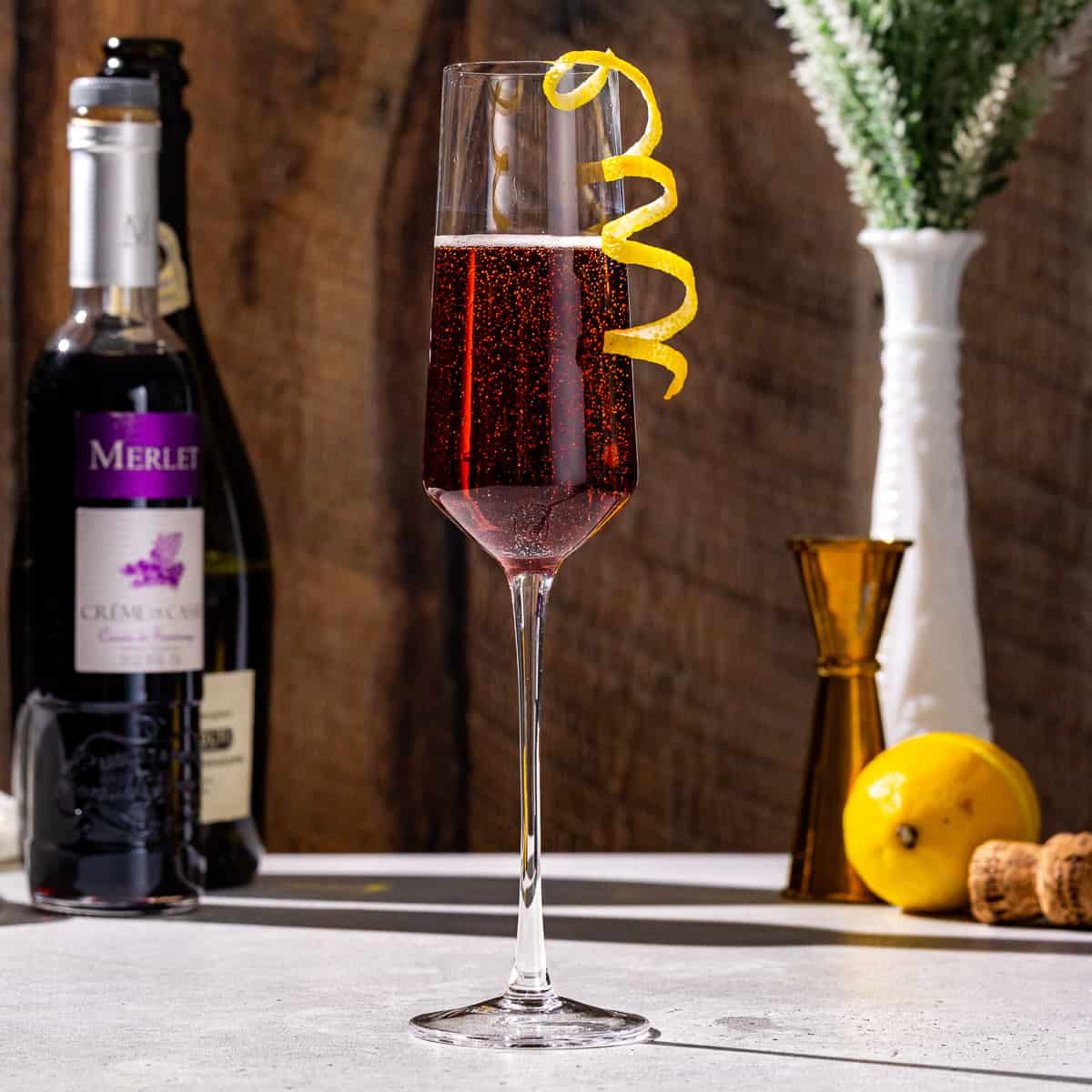 Side view of a Kir Royale cocktail in a champagne flute. A bottle of creme de cassis, a lemon, a jigger, a champagne cork and a bottle of prosecco are in the background.