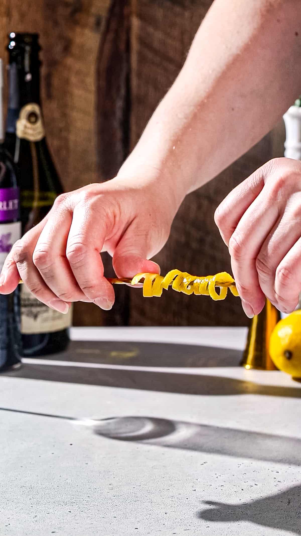 Hands twist a long strip of lemon peel around a bar spoon handle.