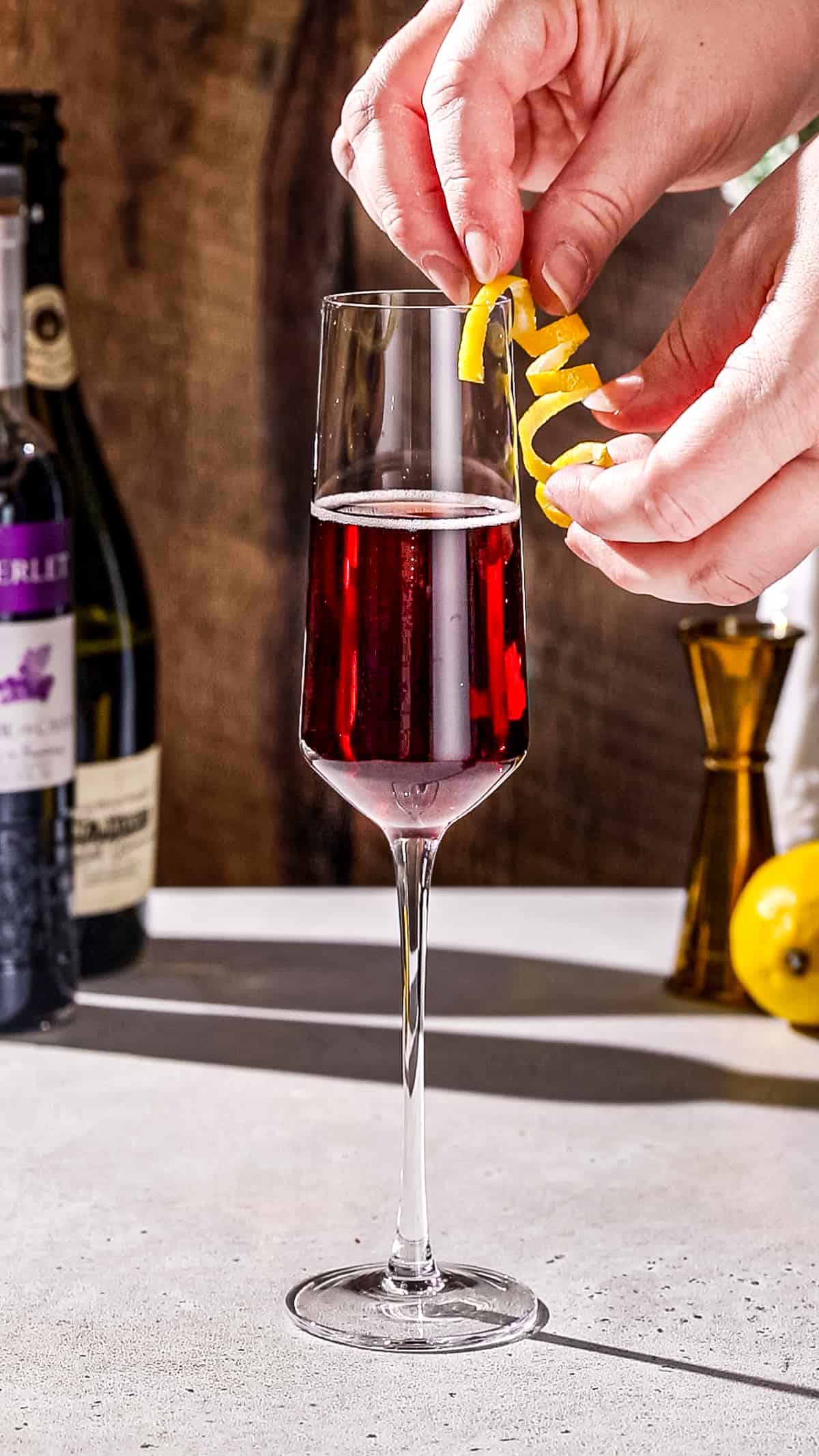 Hand adding a long curly lemon peel garnish to a champagne flute filled with red liquid.