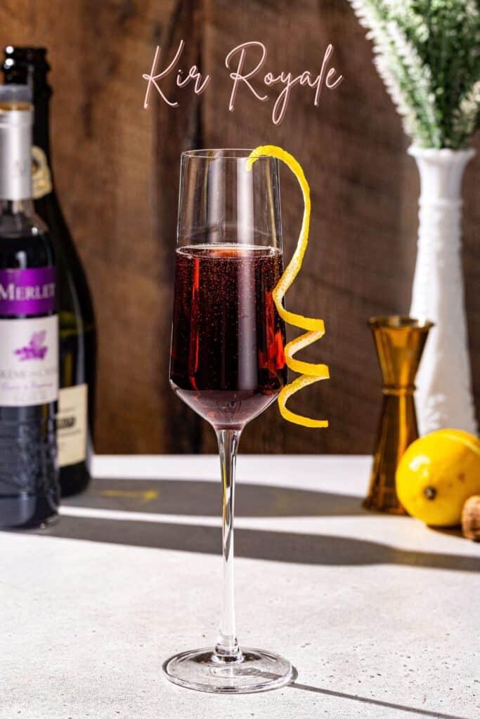 Overhead view of a Kir Royale cocktail in a champagne flute. The drink is fizzy and has a lemon peel garnish. A lemon and a champagne cork are in the background. Script text above the drink says “Kir Royale”.