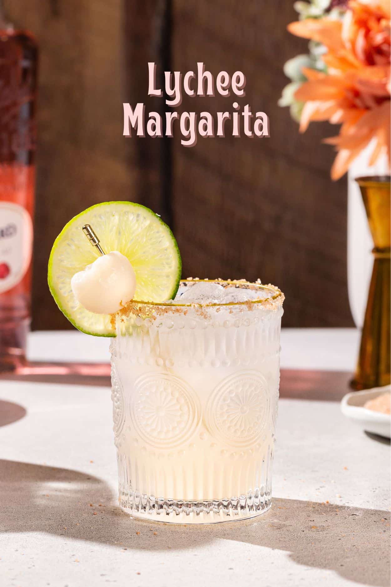 Side view of a Lychee Margarita cocktail with a lime slice garnish and a whole lychee garnish. In the background is a bottle of liqueur and a jigger. Text overlay above the drink says “Lychee Margarita”.