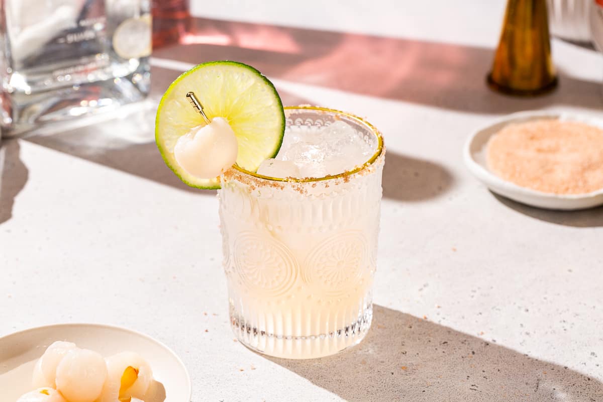 Lychee Margarita cocktail on a countertop. The drink is garnished with a lime and a lychee, as well as a salt rim. A dish of salt and a dish of lychee can be seen around the glass, and there are some bottles and a jigger partially seen in the background.