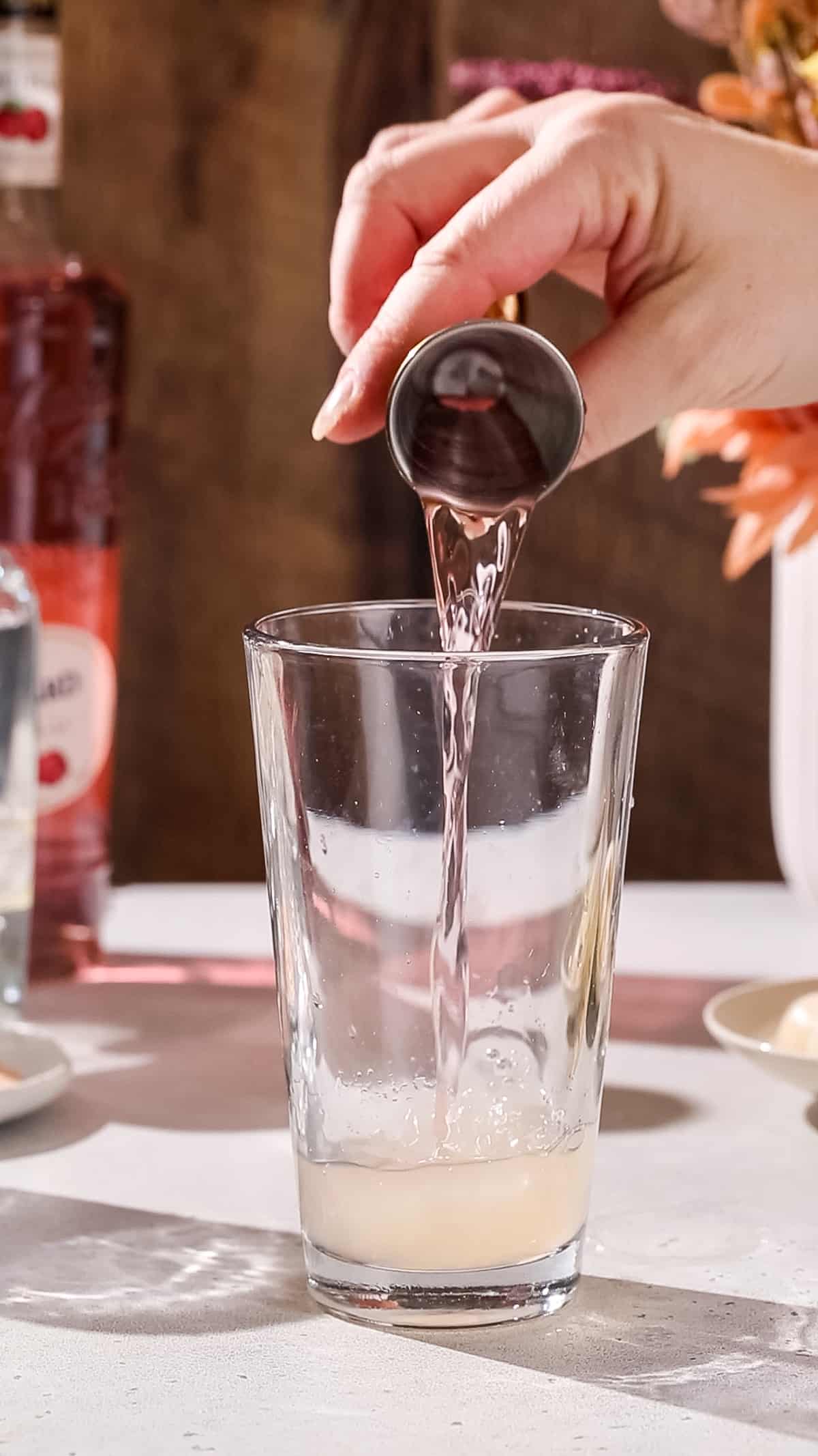 Hand pouring lychee liqueur into a cocktail shaker.