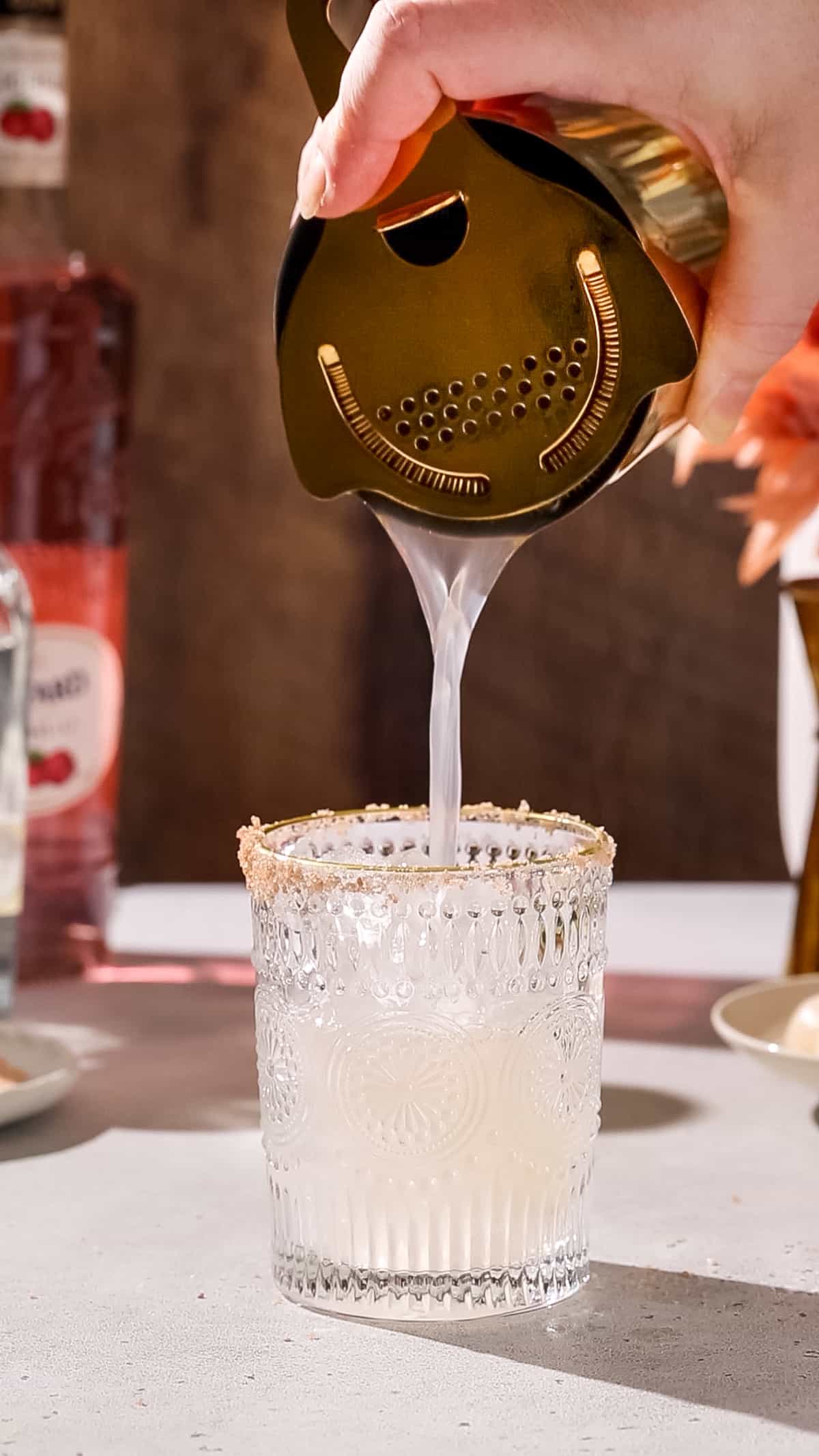 Hand using a cocktail shaker to strain a drink into a prepared glass.