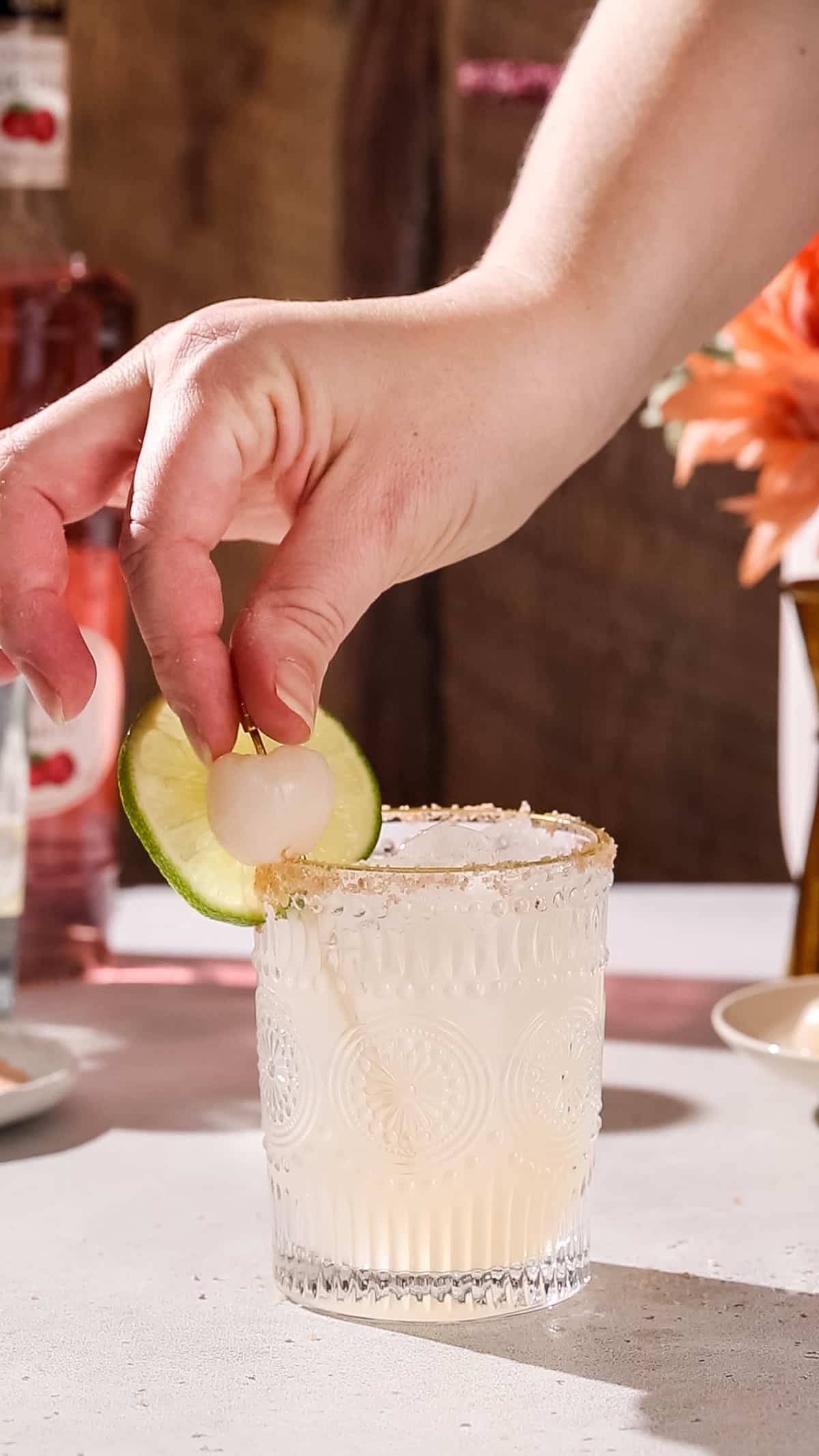 Hand adding a lychee fruit on a cocktail pick to a cocktail glass as a garnish.