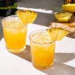 Slightly overhead view of two Pineapple whiskey sour cocktails on a countertop. They are yellow in color and have a pineapple wedge as a garnish in each drink. In the background is a cutting board with some fresh lemon and pineapple, along with a bottle of whiskey.