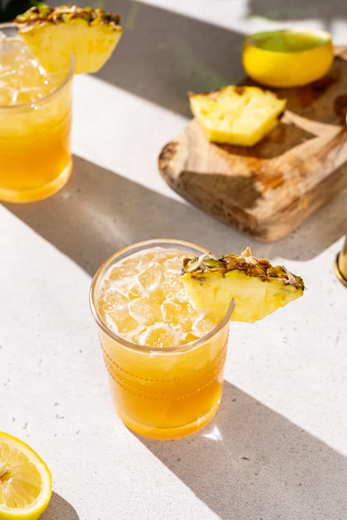 Overhead view of two Pineapple Whiskey Sour cocktails. The drinks are garnished with pineapple wedges and there is fresh pineapple on a cutting board in the background.