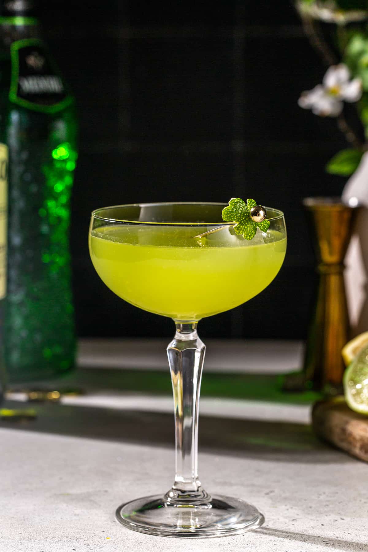 Side view of a green Shamrock Sour cocktail in a coupe glass. The drink is green in color and garnished with a lime peel. In the background are some bottles and bar tools.