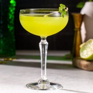 Side view of a green colored Shamrock Sour cocktail on a countertop. A lemon, lime and cocktail jigger are in the background. The drink is garnished with a lime peel cut to look like a shamrock.