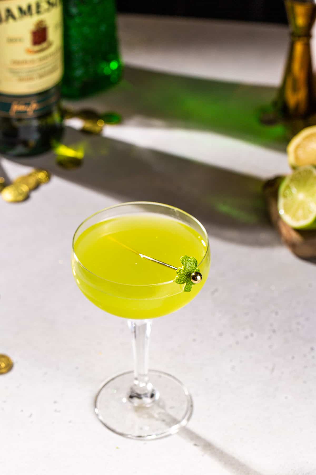 Overhead view of a Shamrock Sour cocktail with a lime peel garnish in the shape of a shamrock. There are cocktail ingredients and bar tools in the backround.