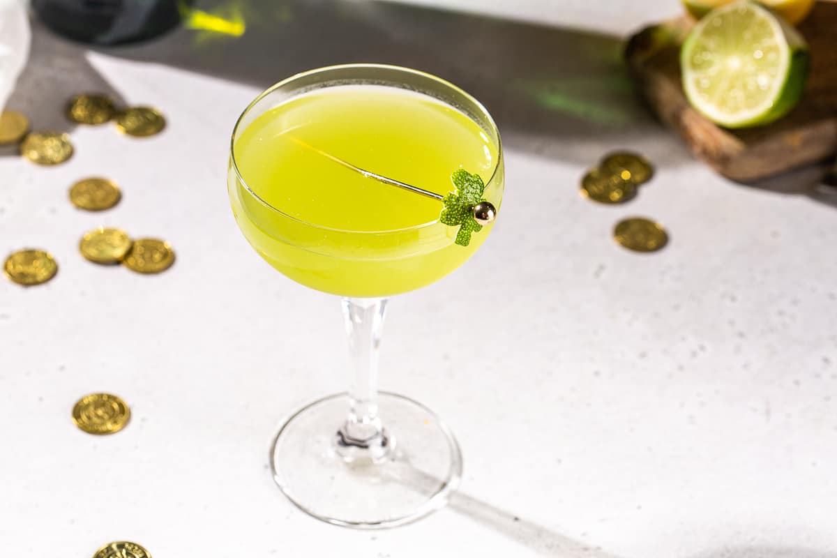 Shamrock Sour cocktail in a coupe glass on a countertop. The drink is green and has a lime peel garnish in the shape of a shamrock. There are some gold coins and a lime in the background.